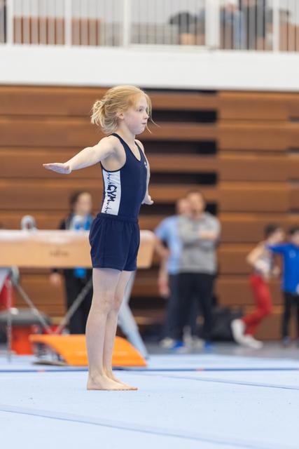 Young gymnast with red hair in navy leotard performs floor routine with arms extended, focused expression at indoor meet