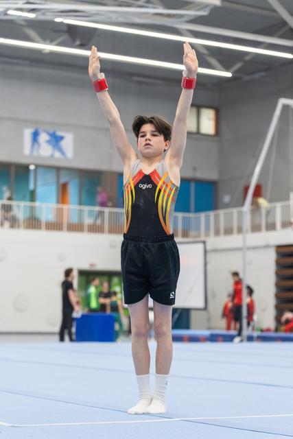 Young gymnast in colorful leotard stands with arms raised overhead on blue floor mat in indoor training facility