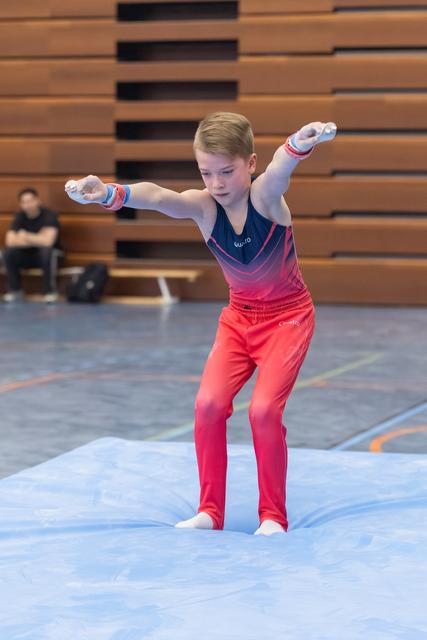 Young male gymnast in gradient pink uniform performs floor routine with arms extended, showing focused concentration