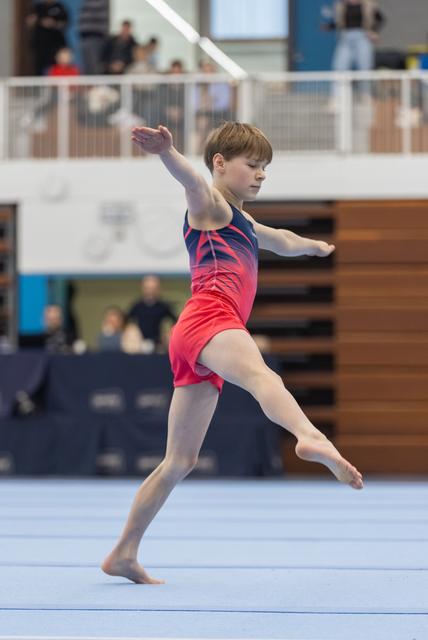 Young gymnast performs an elegant dance element during floor routine, arms extended gracefully in pink and navy leotard