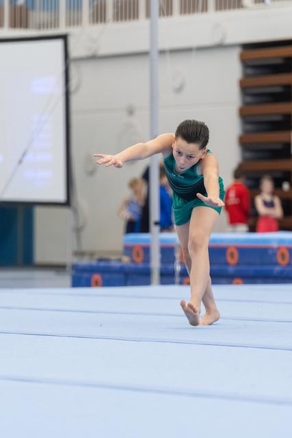 Young gymnast in green leotard performs floor routine with intense focus and extended arm position on blue training mat