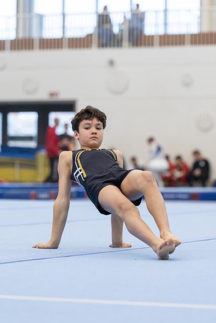 Young gymnast performing floor routine with backward lean position, demonstrating flexibility and control on blue mat