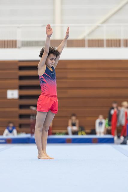 Young gymnast stands with arms raised overhead on floor mat, focused expression, preparing for routine in sports hall