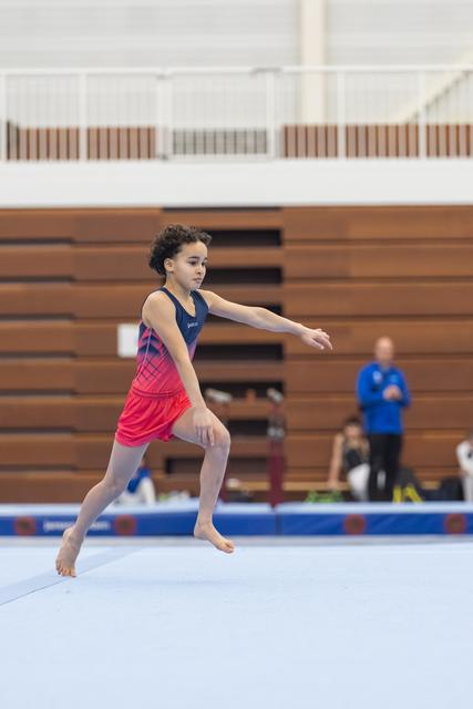 Young gymnast performs floor routine with extended arms, displaying focused concentration in gymnasium with bleachers