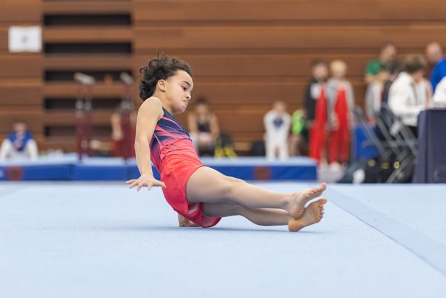Young gymnast performs a seated floor exercise position with legs extended, demonstrating control and form during routine