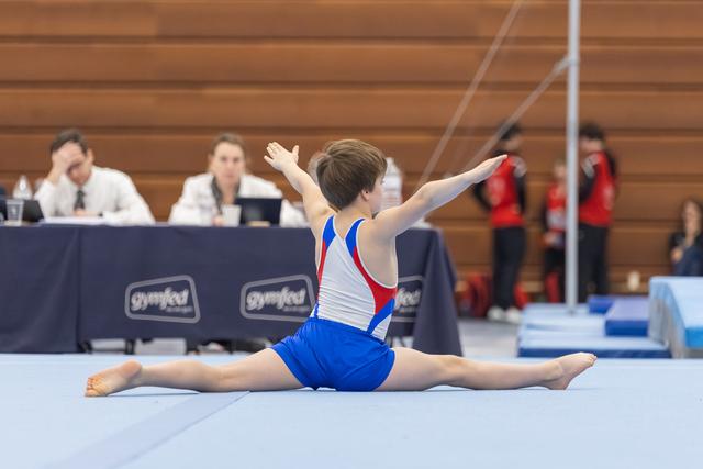 Young gymnast performs full split stretch on floor mat with judges observing in background during gymnastics meet