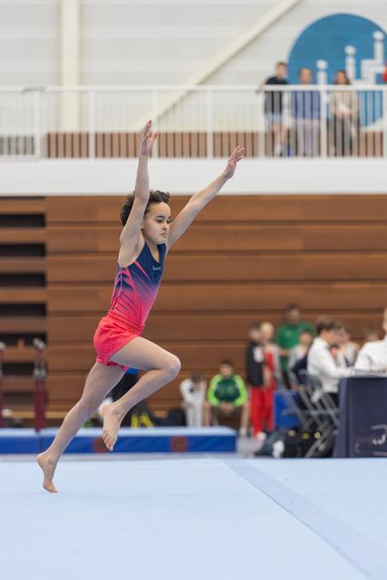 Young gymnast performs graceful floor routine with arms extended upward, wearing vibrant pink-to-blue gradient leotard
