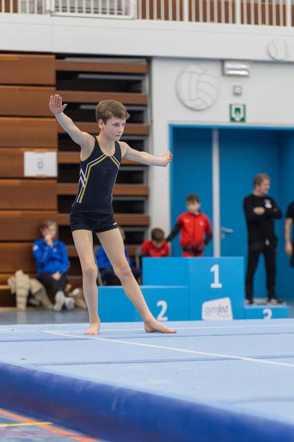 Young gymnast in navy leotard performs floor routine with arms gracefully extended, focused expression in indoor gym