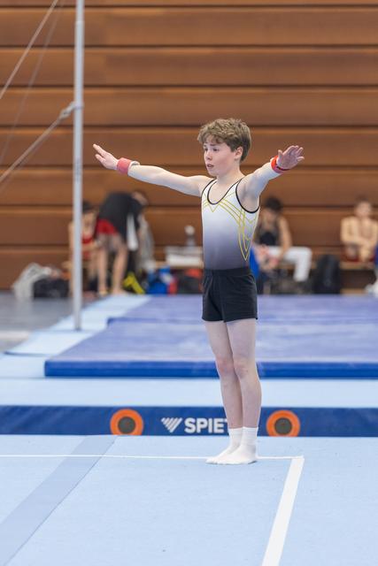 Young male gymnast performing floor routine with arms extended wide, showing focus and determination in gym with wooden walls