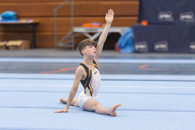 Young gymnast completes floor routine with raised arm, sitting in splits position on blue mat in training facility