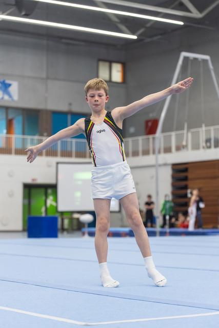 Young gymnast in white uniform performs floor routine with extended arms, displaying focused concentration in sports hall