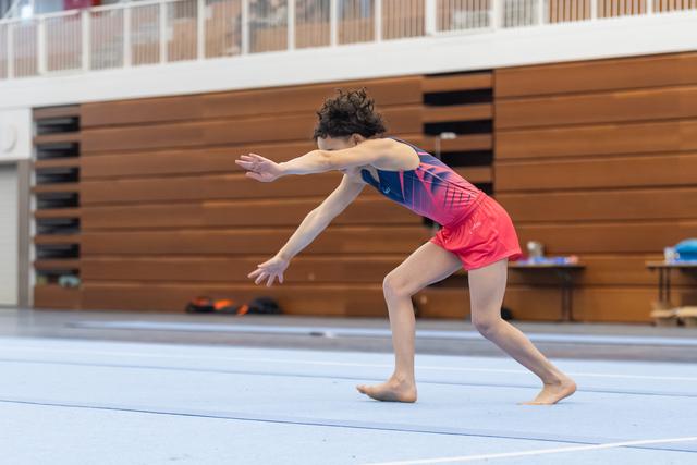 Young gymnast performs floor routine with dramatic forward lunge pose, arms extended, wearing pink and blue leotard in gymnasium