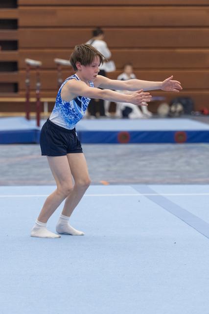 Young gymnast in blue leotard performs expressive arm movements during floor routine, displaying intense focus and concentration