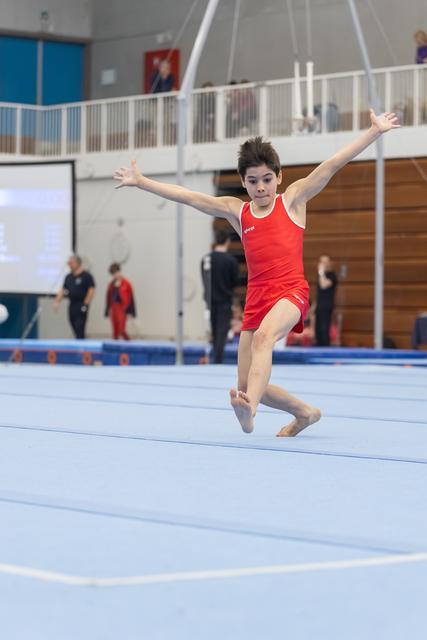 Young gymnast in red leotard performs a leap during floor routine, arms extended, focused expression in training facility