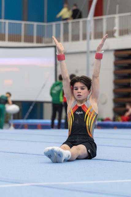Young gymnast in black and yellow leotard sits on floor mat with arms raised overhead, completing a routine element