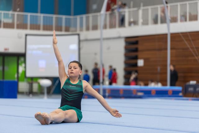 Young gymnast in green leotard performs floor routine with pointed toe and extended arms, showing concentration and grace