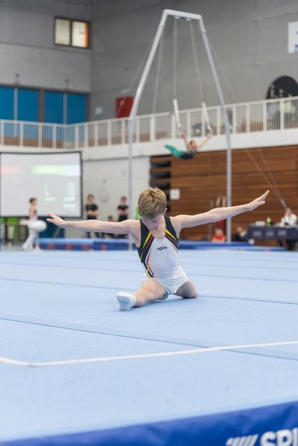 Young male gymnast performs a split position on floor exercise mat with arms extended during routine at indoor facility