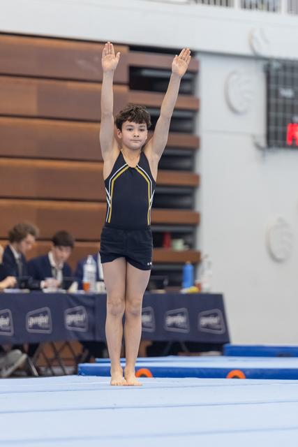 Young gymnast in navy leotard stands on floor mat with arms raised overhead in competition salute, judges table visible behind
