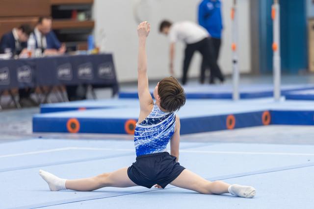 Young gymnast performs floor split routine in blue sequined leotard with arm extended upward during training session