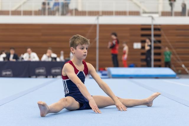 Young gymnast in navy and burgundy leotard performs straddle stretch on floor mat, focused expression during warm-up