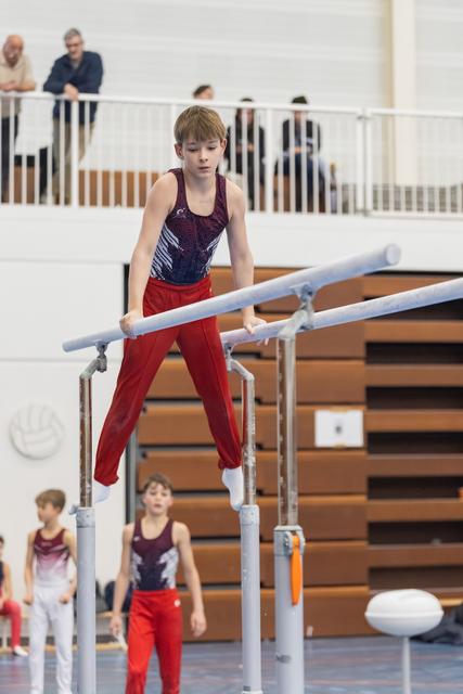 Young gymnast in red pants and purple leotard performs on parallel bars with focused concentration while teammates watch below