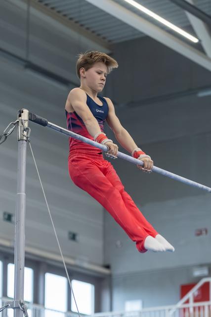 Young gymnast performing on horizontal bar with intense concentration, wearing red pants and navy leotard in training facility