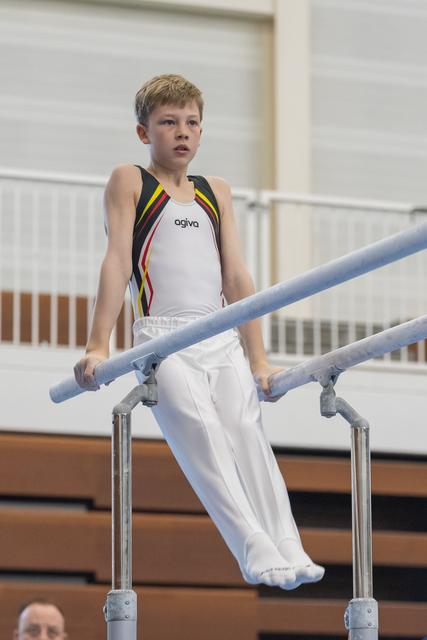 Young gymnast in white and rainbow-striped leotard performs on parallel bars with concentrated expression in gymnasium