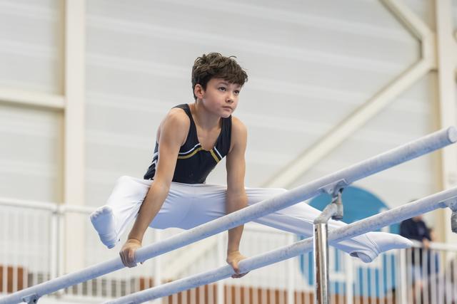Young gymnast gripping parallel bars with focused expression, wearing navy leotard and white pants in indoor training facility