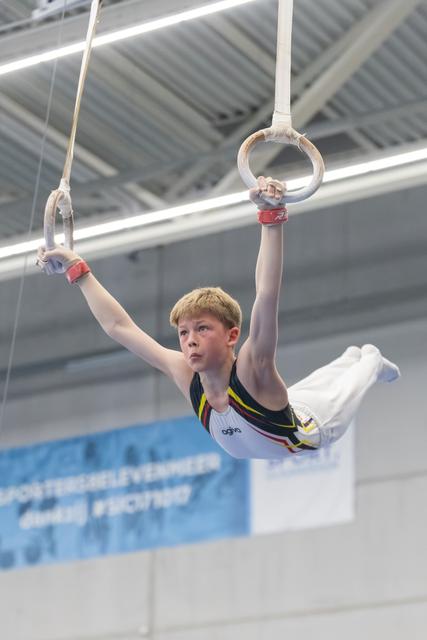 Young male gymnast performing on rings, displaying intense concentration while holding position in indoor training facility