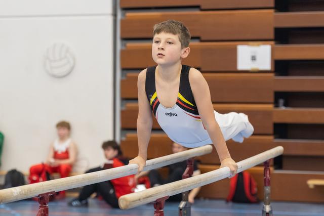 Young gymnast performs on parallel bars with intense concentration, while teammates in red wait in background