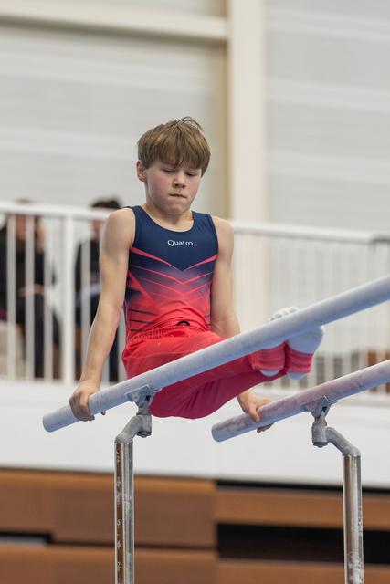Young gymnast in red gradient leotard concentrating intensely while performing on parallel bars at indoor training facility