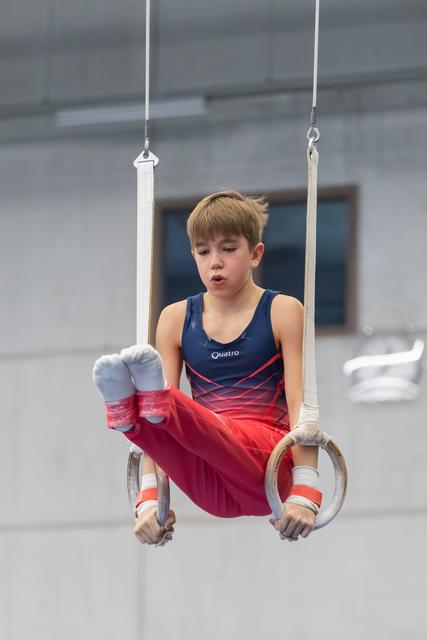 Young gymnast in blue and red leotard performing on rings with intense concentration, legs extended horizontally