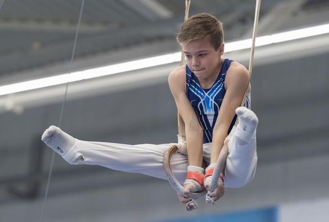Young male gymnast performing on rings with concentrated expression, legs extended in white pants, blue leotard visible
