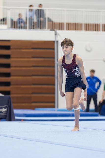 Young gymnast in sequined leotard walks across floor mat with focused expression during routine at gymnastics meet