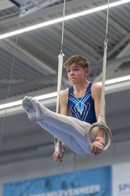 Young male gymnast performing an L-sit hold on still rings, displaying intense concentration during his routine