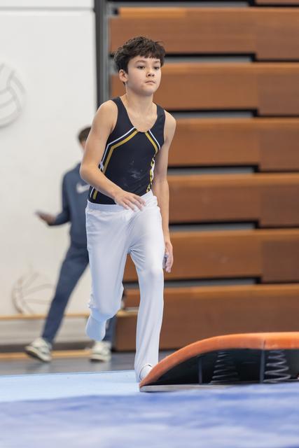 Young gymnast in navy leotard and white pants walks with focused expression on blue floor mat, wooden bleachers behind