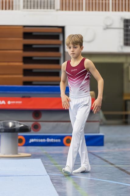 Young gymnast in burgundy and white leotard walks with focused expression across training floor at gymnastics facility