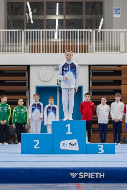 Young gymnast stands proudly on first place podium wearing blue and white tracksuit, with competitors and teams lined up behind