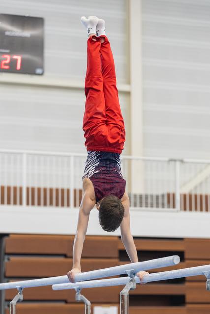 Young gymnast performs a handstand on parallel bars during training, wearing red hoodie and purple shorts in indoor gym