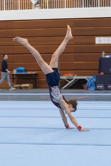 Young gymnast performing a handstand on blue floor mat, legs extended upward, in training facility