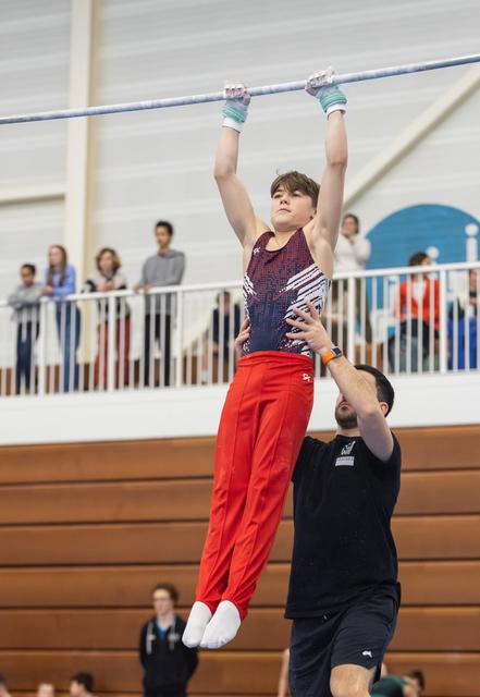 Young gymnast in red pants and sparkly leotard hangs from high bar while coach spots from below in indoor gymnasium