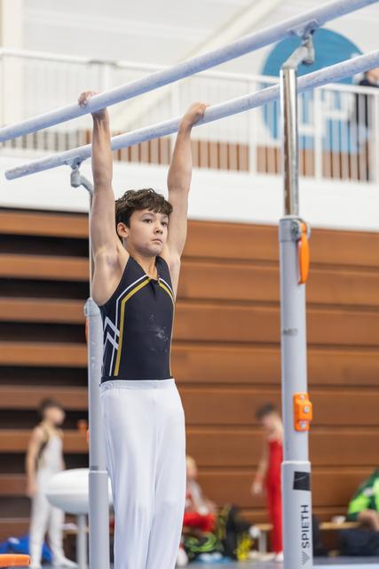Young gymnast in navy and yellow leotard hanging from horizontal bar with focused expression during indoor routine