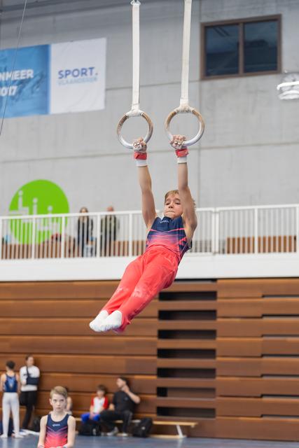 Young gymnast in red pants and striped leotard hangs from still rings, body extended, during training session in gymnasium
