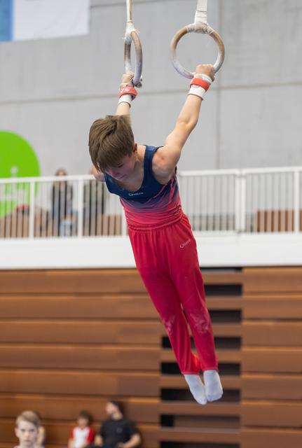 Young gymnast in pink pants and blue leotard hanging from gymnastics rings during practice in an indoor gym facility