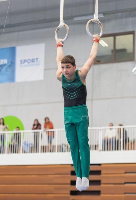 Young gymnast in teal uniform hangs from rings with focused expression during indoor gymnastics event
