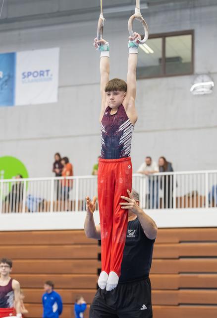 Young gymnast in red pants hangs from rings with focused expression while coach spots from below in indoor gym