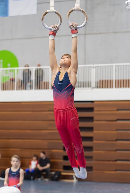Young gymnast in gradient pink outfit hangs from rings with focused expression during training session in indoor gymnasium