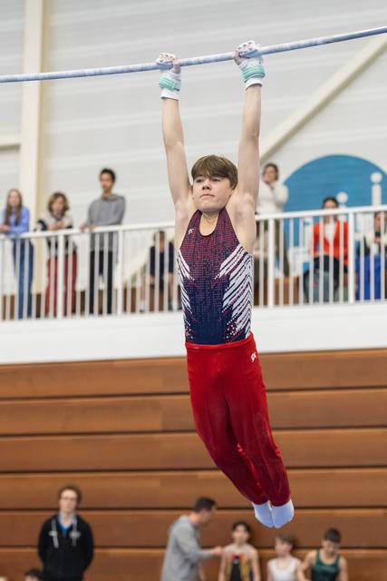 Young gymnast in sparkly leotard and red pants hangs from horizontal bar, arms extended overhead, spectators watching from balcony