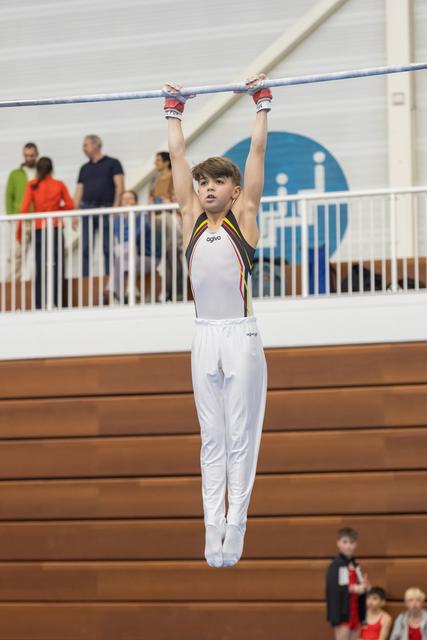 Young gymnast in white uniform hangs from horizontal bar with determined expression, spectators watching from gallery above