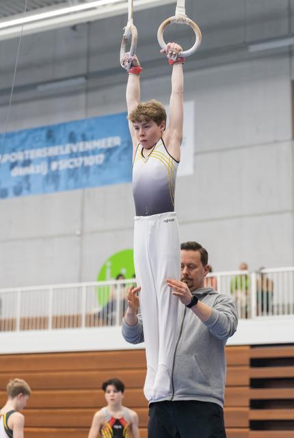 Young gymnast performs a hang on rings while his coach spots him closely, hands ready to assist in the training facility
